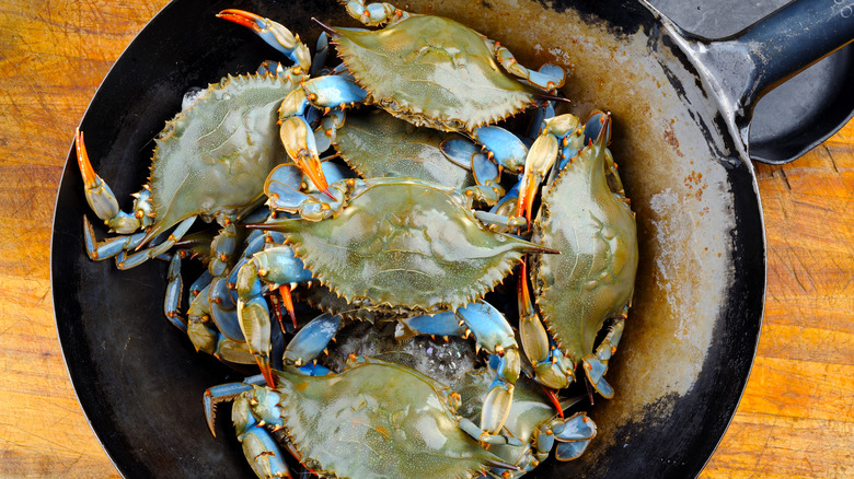 Several blue crabs are piled inside a weathered black wok on a well0worn wooden tabletop.