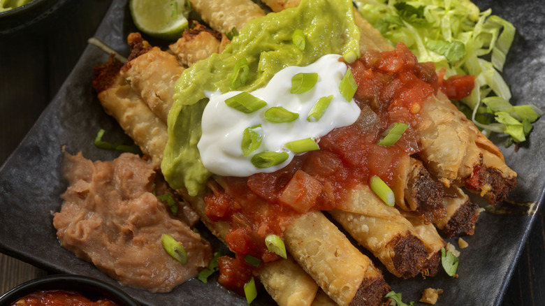 a taquito platter topped with salsa, guacamole, sour cream, and green onions served with a side of lettuce and refried beans