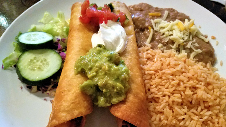 a plate of flautas topped with guacamole, sour cream and salsa surrounded by rice, refried pintos and a salad