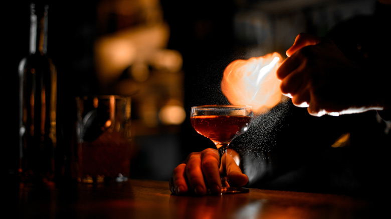 Bartender's hands holding a glass with a cocktail and spraying it with flame spray