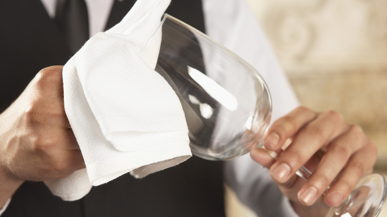 A person in a black vest, white long-sleeved shirt, and black tie polishes a wine glass with a cloth held in their two hands.