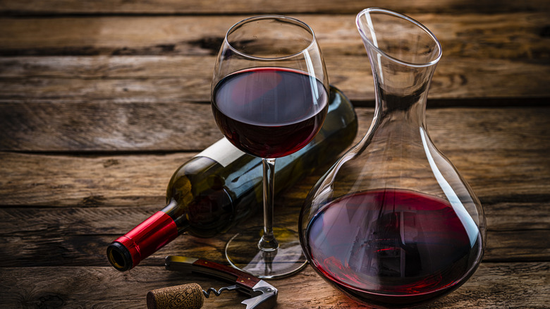 A wine decanter filled with dark red wine, a tall wine glass also filled with wine, a corkscrew, and a side-laying wine bottle with a red band are all arranged on a rustic wooden surface.