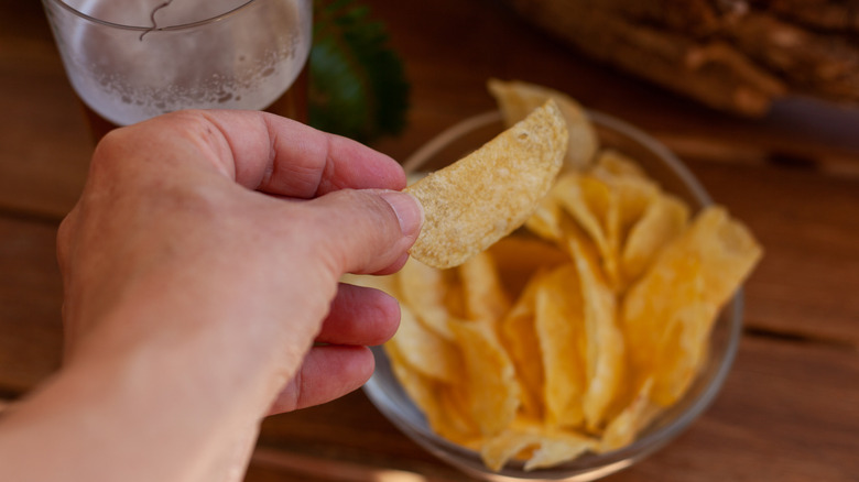 Hand reaching into a bowl of potato chips with a beer in the background on a wooden table.