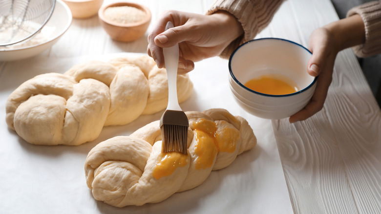 Hands brushing egg wash onto two risen loaves of challah.